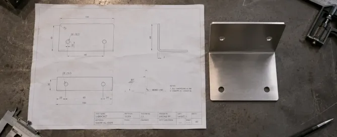 Eine gedruckte 2D-Technikzeichnung einer Blechhalterung neben dem gefertigten Metallteil auf einer Werkbank