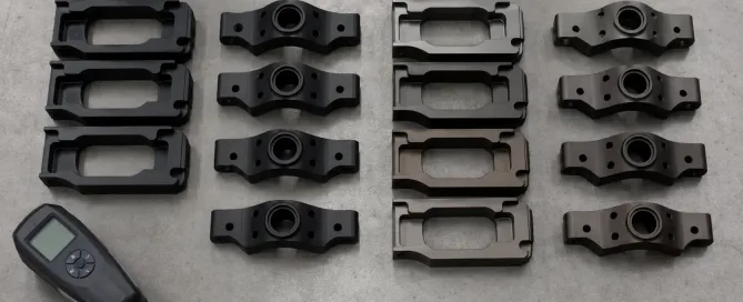 Schwarz eloxierte Aluminiumteile auf einer Prüffläche neben Farbabweichungsmustern angeordnet, die inkonsistente schwarze Töne zeigen